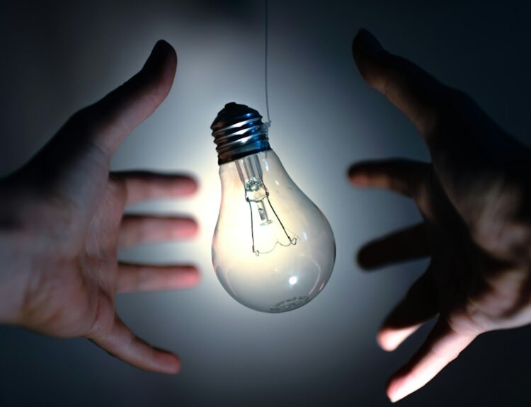 Two hands reaching towards a light bulb in a dark setting, symbolizing ideas or innovation.