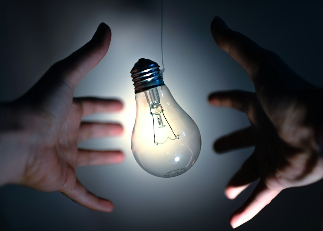 Two hands reaching towards a light bulb in a dark setting, symbolizing ideas or innovation.